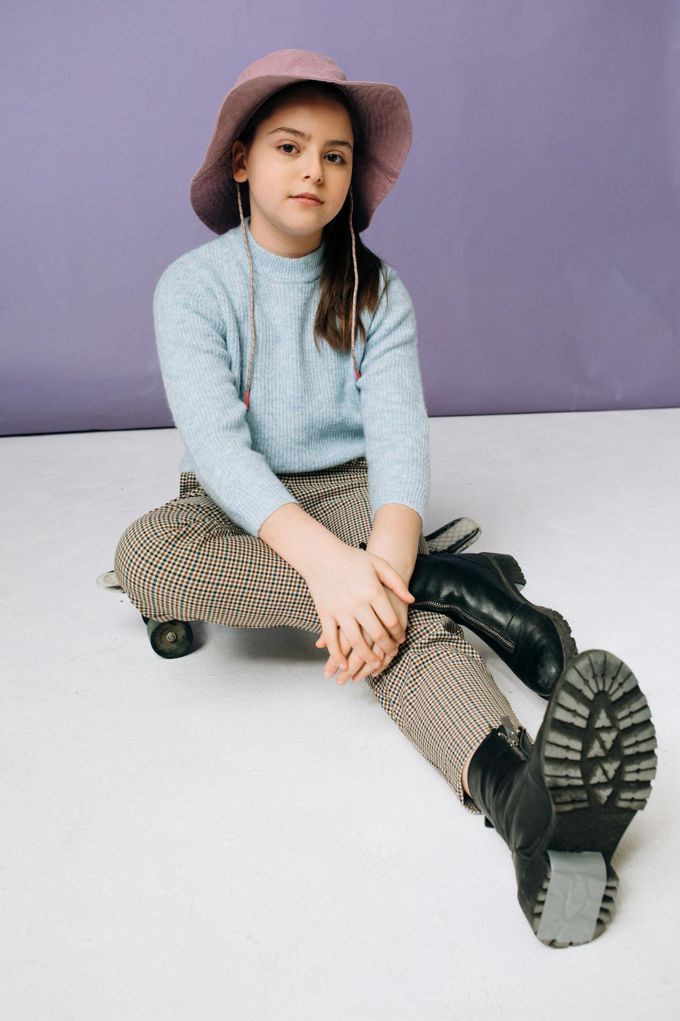 Portrait of a young girl in a stylish outfit with a purple hat, sitting against a pastel background.